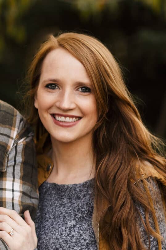 A young woman with long auburn hair smiles at the camera. She is wearing a grey sweater and a brown jacket. The blurred greenery in the background hints at a peaceful day near Minnesota’s Rainy Lake. | Rainy Lake, MN