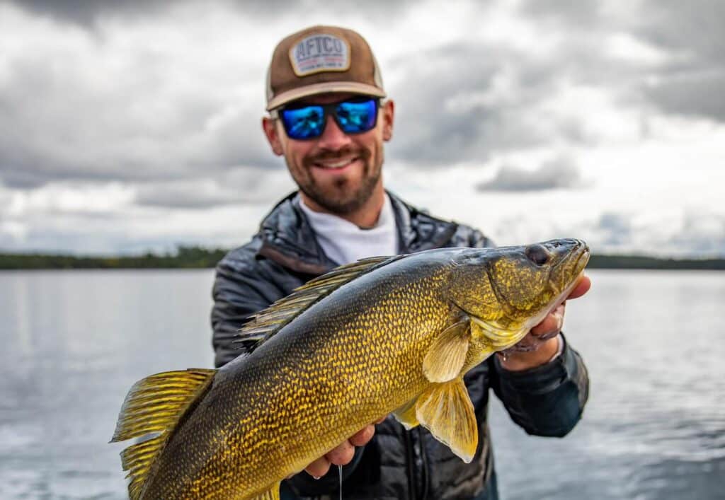 A smiling man wearing sunglasses and a cap holds a large, golden fish toward the camera, standing on a boat with Rainy Lake and a cloudy sky in the background. | Rainy Lake, MN