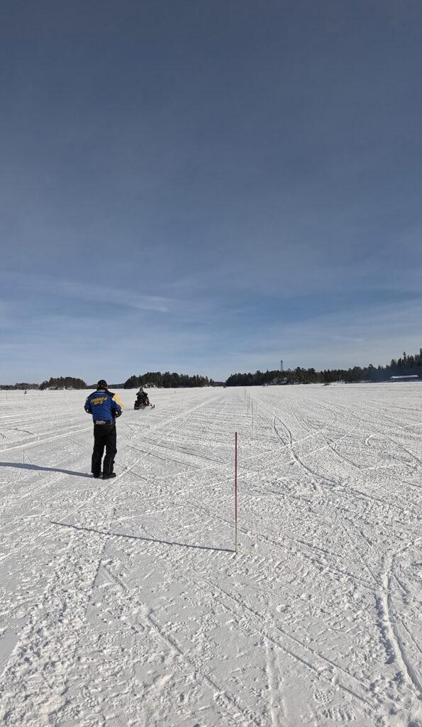 A person in winter clothing walks across a snowy, frozen Rainy Lake in Minnesota toward a distant snowmobile under a clear blue sky, with a red pole marking the snow and trees visible in the background. | Rainy Lake, MN