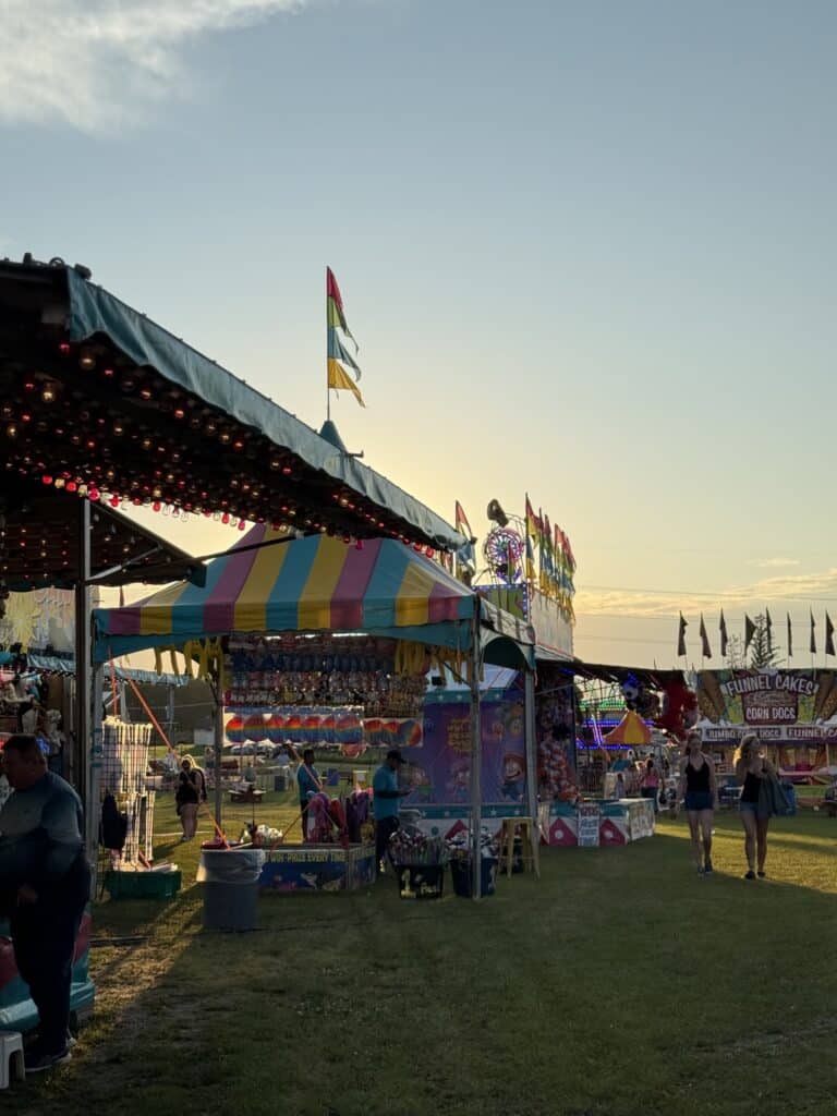 Brightly colored carnival booths and tents with games and food stalls are set up outdoors near Rainy Lake. People walk along the grassy path as the sun sets, creating a warm, festive atmosphere. Flags and lights decorate the lively Minnesota scene. | Rainy Lake, MN