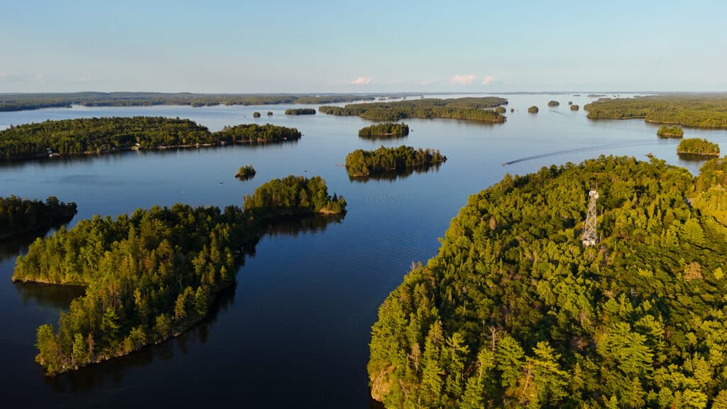Aerial view of Minnesota’s Rainy Lake with numerous tree-covered islands and calm blue water. A boat creates a wake, while a tall observation tower rises among the trees on one of the islands. | Rainy Lake, MN