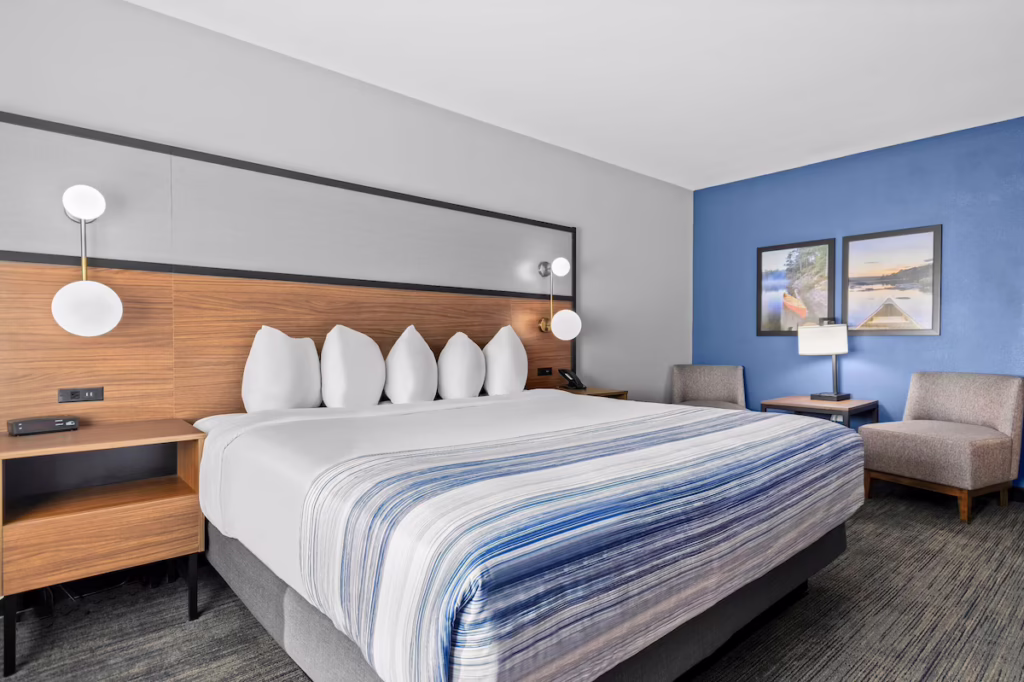A modern hotel room in Minnesota with a king-sized bed, striped bedding, five pillows, wooden headboard, bedside tables, two wall lamps, chairs, a small table, and framed pictures on a blue accent wall—perfect after exploring Rainy Lake. | Rainy Lake, MN