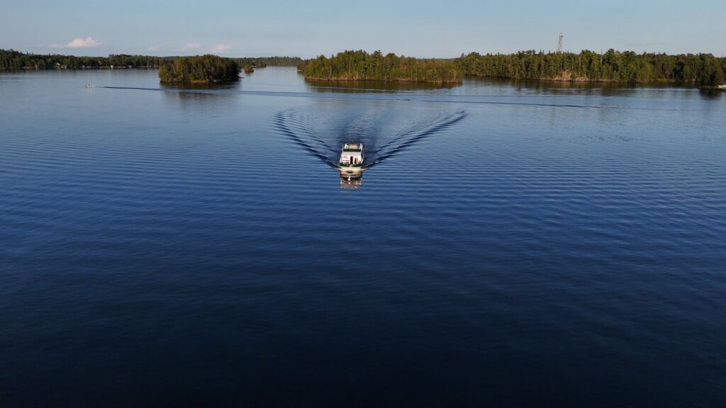 A small boat moves across Minnesota’s calm Rainy Lake, leaving ripples behind it. The boat heads toward tree-covered islands under a clear sky. | Rainy Lake, MN