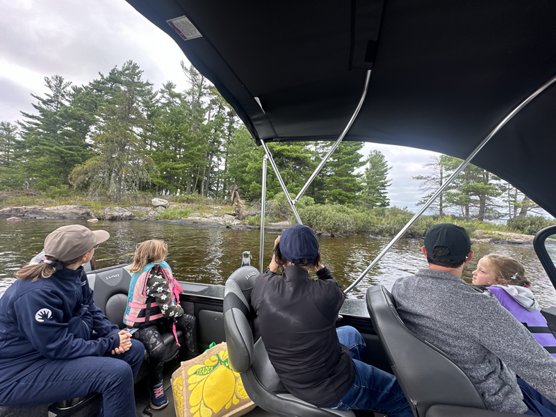 Four adults and two children sit in a boat on a lake during one of the park tours at Voyageurs National Park, looking toward a forested shoreline under a cloudy sky. One person is taking a photo as the group relaxes and enjoys the scenic view. | Rainy Lake, MN