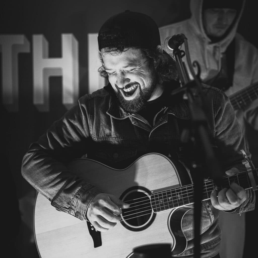 A bearded man wearing a cap and denim jacket smiles widely while playing an acoustic guitar and singing into a microphone. Another musician is visible in the background. The image is in black and white. | Rainy Lake, MN