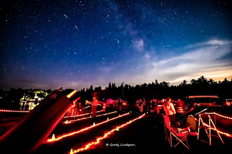A group of people gather outdoors at night with telescopes under a star-filled sky, illuminated by red lights on the ground and surrounded by trees. Some meteor trails are visible above. | Rainy Lake, MN