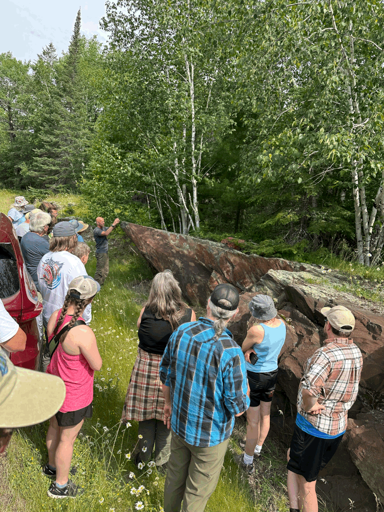 A group of people stands outdoors by a large, slanted rock formation, listening to a guide who is gesturing toward the rocks. They are surrounded by green trees and grass on a sunny day. | Rainy Lake, MN
