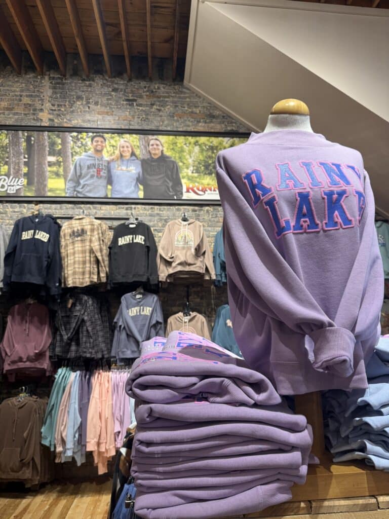 A clothing store display featuring folded and hanging Rainy Lake Gear sweatshirts in various colors, with a mannequin wearing a lavender version. A large photo of three people in gear is on the wall above. | Rainy Lake, MN