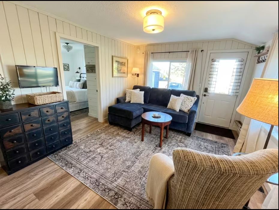 A cozy living room with a fir-toned dark sectional sofa, round wooden coffee table, patterned area rug, armchair, TV on a drawer chest, and bright windows by the entrance door. A thistle-adorned bedroom is visible through an open doorway. | Rainy Lake, MN