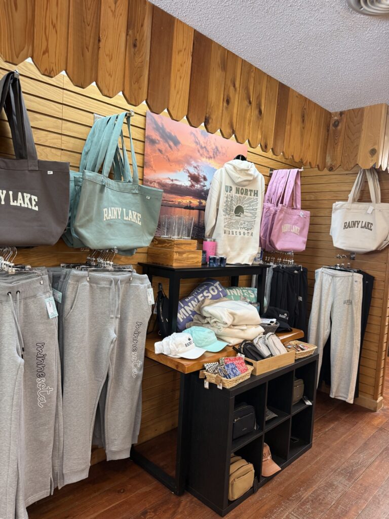 A clothing store display featuring Rainy Lake Gear—sweatshirts, sweatpants, tote bags, hats, and pouches with Minnesota and Rainy Lake branding—arranged on shelves and hooks against a wood-paneled wall with a lake photo above. | Rainy Lake, MN