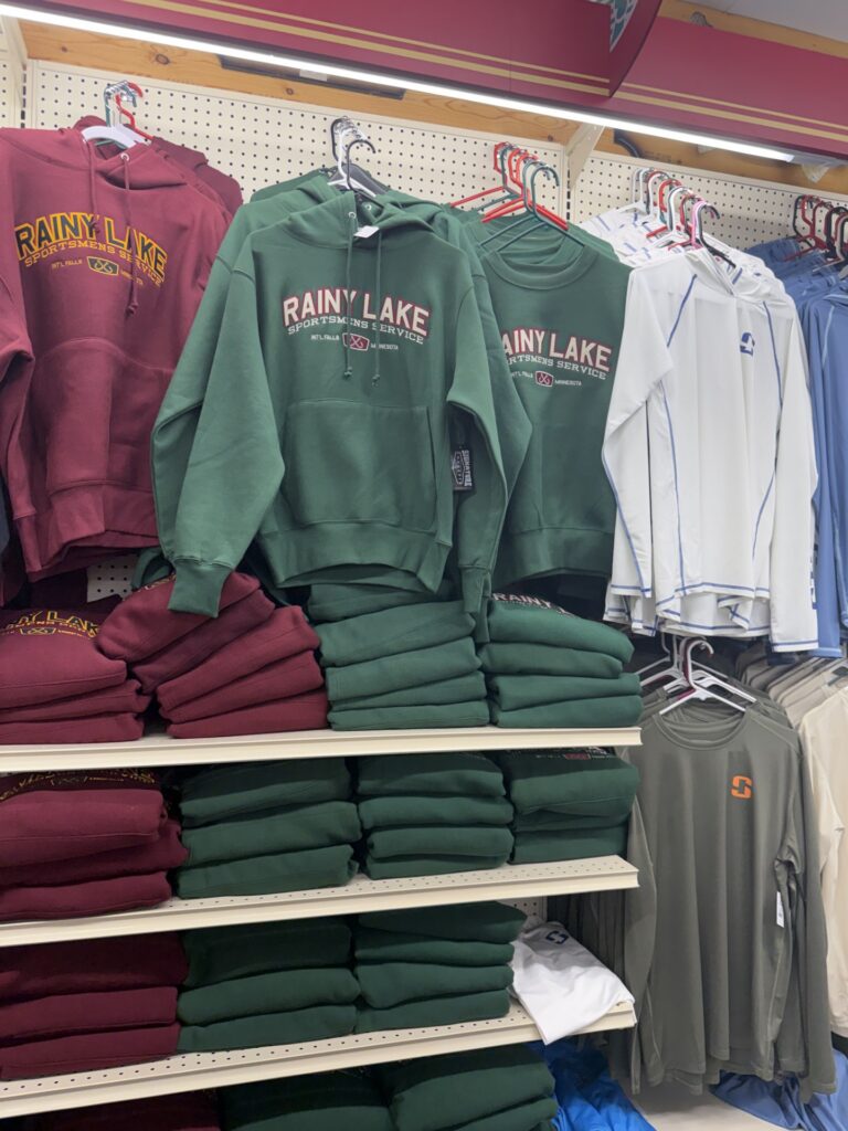 A store display of stacked green and maroon "Rainy Lake" hoodies on shelves, with matching sweatshirts hanging above. On the right, light gray and olive shirts showcase even more Rainy Lake gear on racks. | Rainy Lake, MN