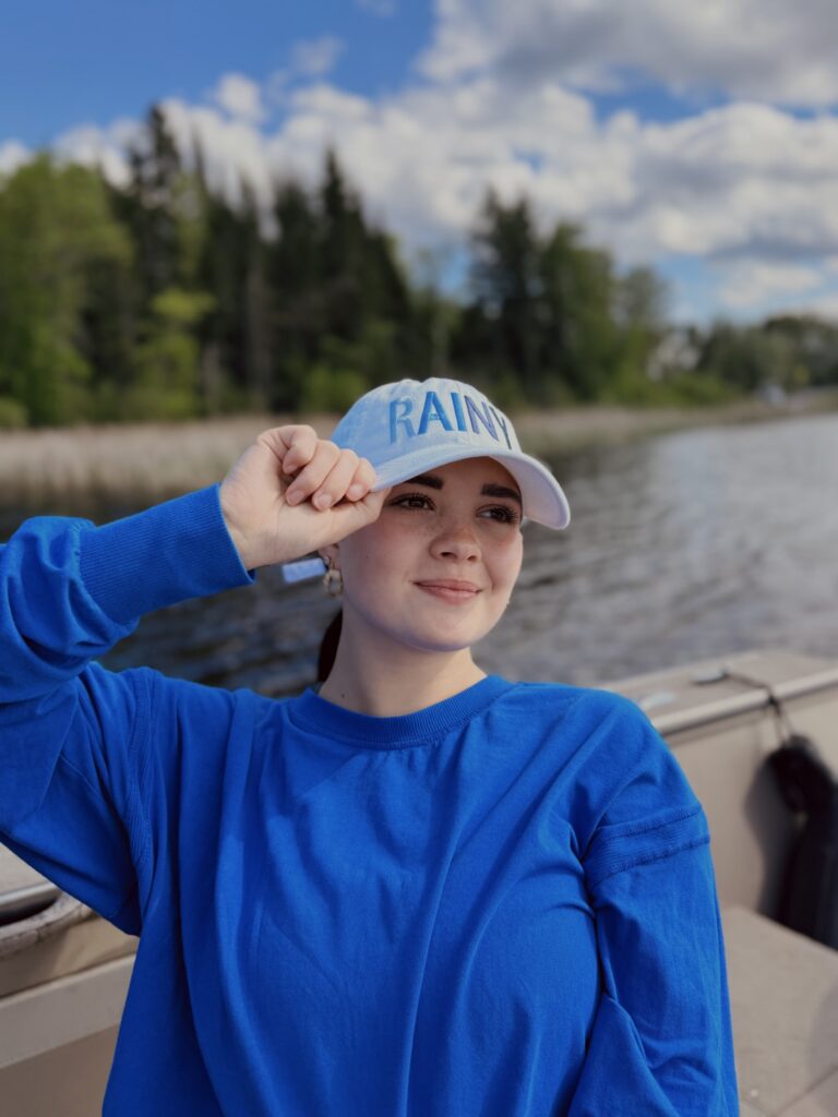 A woman wearing a blue sweater and light blue "RAINY" cap sits on a boat, smiling with one hand holding the brim of her hat