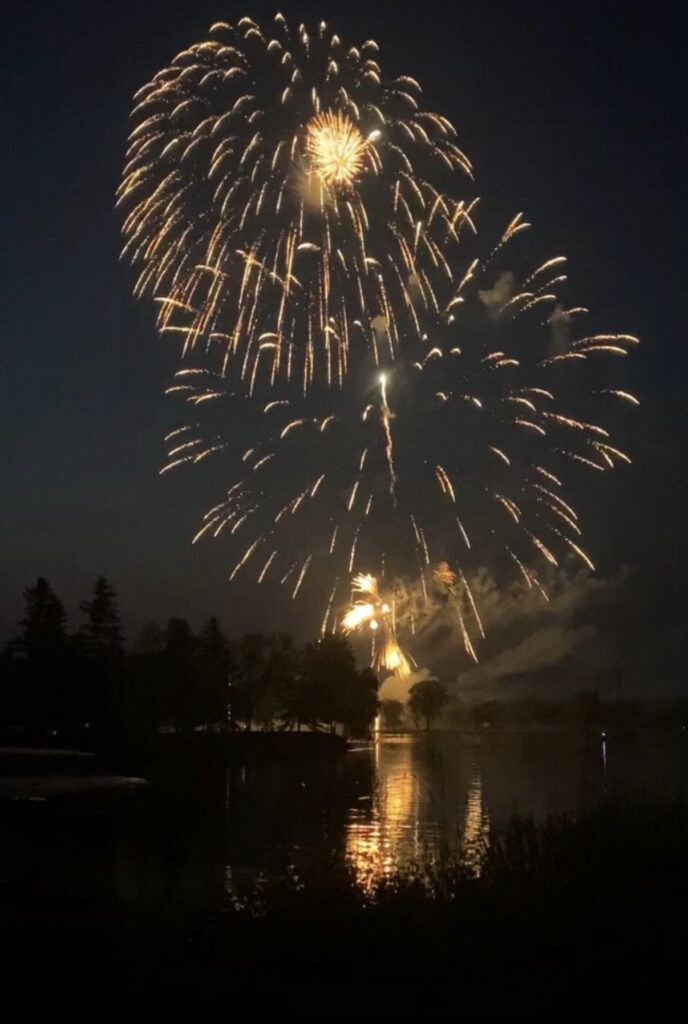 Golden fireworks burst in the night sky over a lake on the 4th of July, reflecting brightly on the water below. Trees and a dark shoreline are silhouetted in the foreground, capturing the spirit of family fun. | Rainy Lake, MN