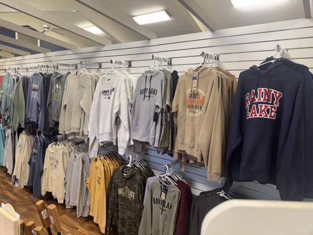 A store display with various hoodies, sweatshirts, and t-shirts—Rainy Lake gear—hanging on a white slat wall. Most clothing features “Rainy Lake” printed logos in assorted colors and styles. The floor is wood-patterned. | Rainy Lake, MN