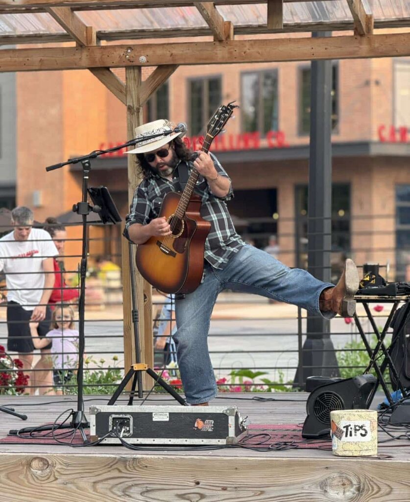 A musician in a cowboy hat and sunglasses plays acoustic guitar on an outdoor stage in International Falls, lifting one leg as he performs. With a tips box up front, he entertains a small crowd during the lively 4th of July festivities. | Rainy Lake, MN