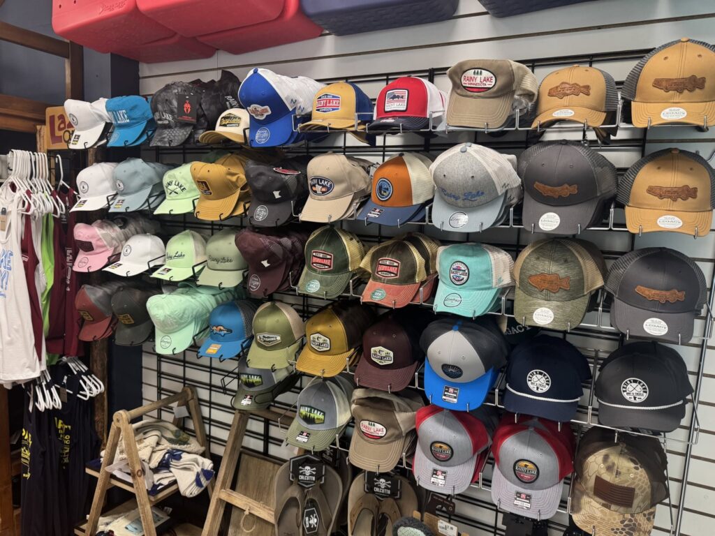 A wall display of colorful baseball caps, including Rainy Lake gear with assorted logos and designs, is neatly arranged on hooks above wooden ladders and racks with T-shirts in this inviting retail store. | Rainy Lake, MN