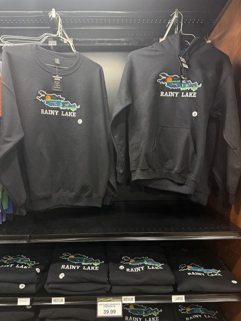 Black sweatshirts and hoodies with a colorful embroidered Rainy Lake design hang on a rack above folded Rainy Lake gear, all displayed for sale. A price tag shows $39.99, with various sizes available. | Rainy Lake, MN