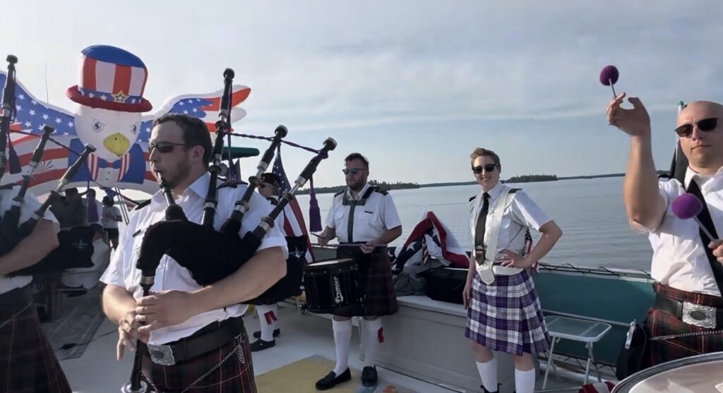 A group of people in kilts play bagpipes and percussion on a boat decorated with American flags and a large inflatable bald eagle, celebrating Independence with family fun, as water and sky stretch in the background. | Rainy Lake, MN