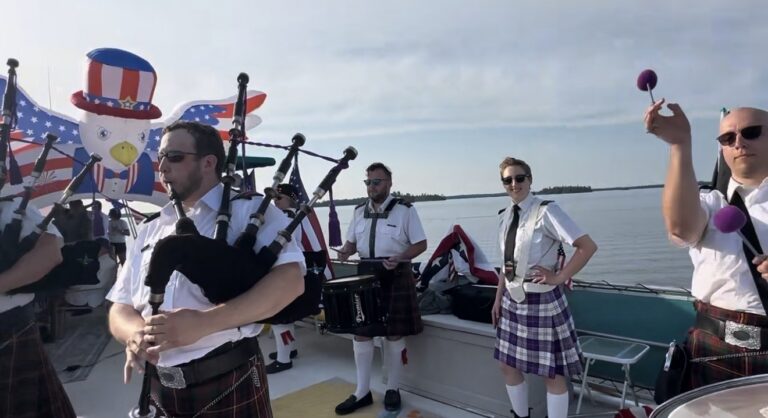 A group of people in kilts play bagpipes and percussion on a boat decorated with American flags and a large inflatable bald eagle, celebrating Independence with family fun, as water and sky stretch in the background. | Rainy Lake, MN