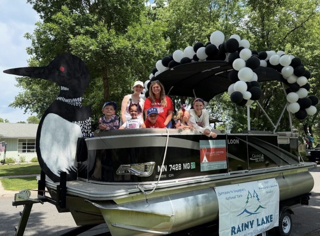 A group of smiling people, including children and adults, stand on a decorated pontoon boat with a large loon figure and black-and-white balloons, celebrating family fun. A “Rainy Lake” banner hangs on the side. Trees and a house are in the background. | Rainy Lake, MN