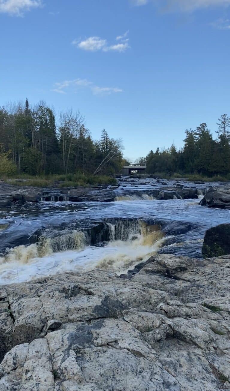 A rocky river with small waterfalls flows through a forested area under a clear blue sky, inviting those chasing waterfalls to explore. Trees line both sides and a bridge is visible in the distance, reminiscent of waterfalls near International Falls. | Rainy Lake, MN