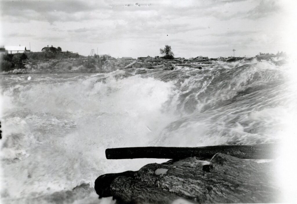 Rushing river water cascades over rocks under a cloudy sky, with a log in the foreground and distant buildings and trees on the horizon—one of the beautiful waterfalls near International Falls MN. | Rainy Lake, MN