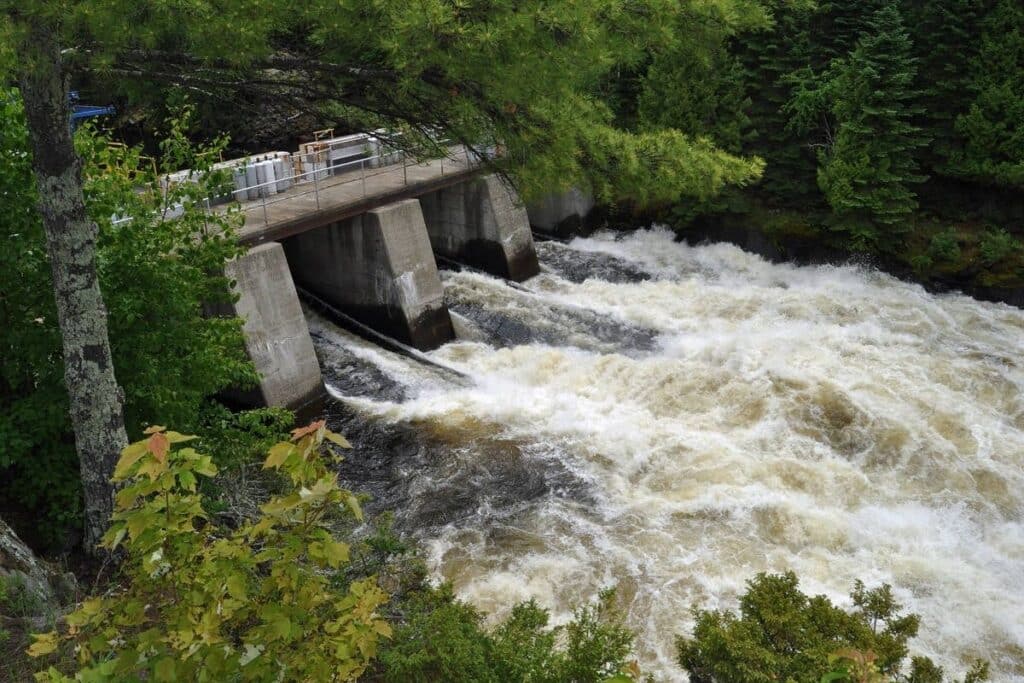 A concrete dam releases rushing white water into a rocky river, surrounded by dense green forest near International Falls MN. The turbulent water flows powerfully, perfect for those Chasing Waterfalls or seeking waterfalls near International Falls MN. | Rainy Lake, MN