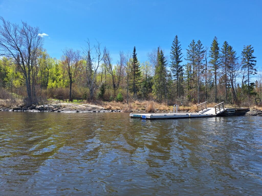 A wooden dock extends into a calm lake, surrounded by trees with fresh green foliage on a sunny day. The shoreline has rocks and sparse grass, with a clear blue sky overhead. | Rainy Lake, MN