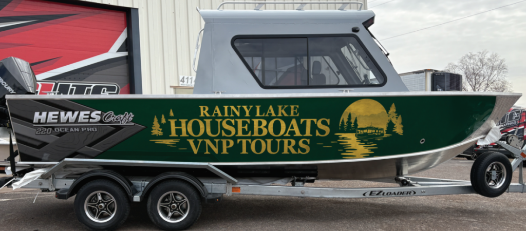 A silver and green HewesCraft 220 Ocean Pro, featuring a "Rainy Lake Houseboats VNP Tours" lake scene logo on the side, is parked outside a building—perfect for exploring Rainy Lake. | Rainy Lake, MN