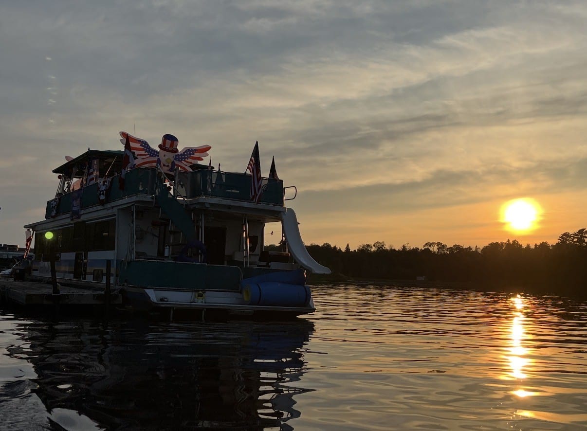 A boat decorated with patriotic American flags and an Uncle Sam inflatable floats on calm water at sunset, capturing the spirit of 4th of July family fun, as trees are silhouetted against the sky and the sun sets on a perfect celebration. | Rainy Lake, MN