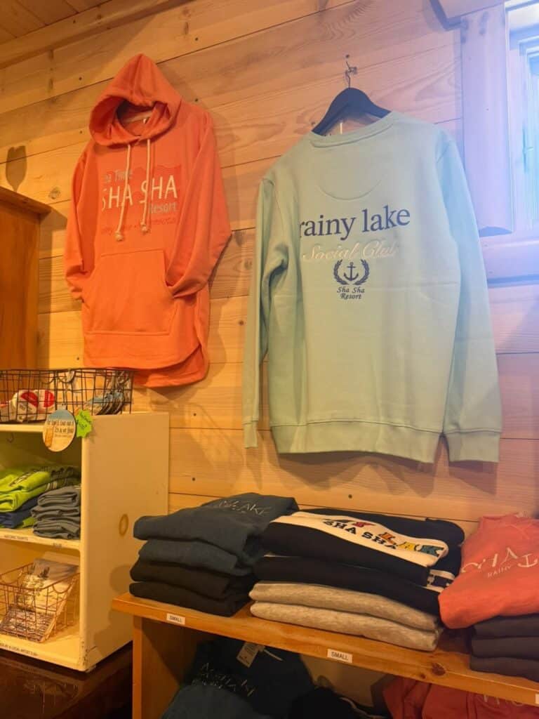 A wooden wall displays a coral hoodie reading "SHA SHA resort" and a light blue "rainy lake Social Club" sweatshirt—perfect Rainy Lake gear for anyone wondering where to find unique apparel. Shelves below hold folded shirts; sunlight streams through a window above. | Rainy Lake, MN