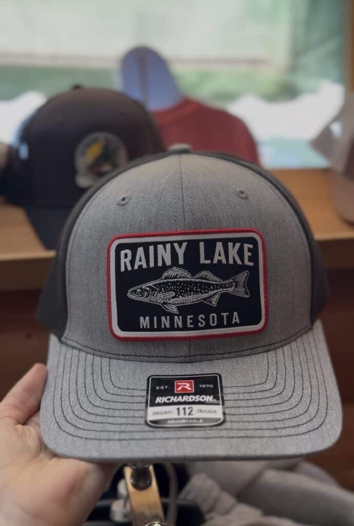 A gray trucker hat with a patch that reads "Rainy Lake Minnesota" and features a fish graphic—perfect Rainy Lake gear. The hat is held in someone's hand, with other hats and outdoor equipment blurred in the background. | Rainy Lake, MN
