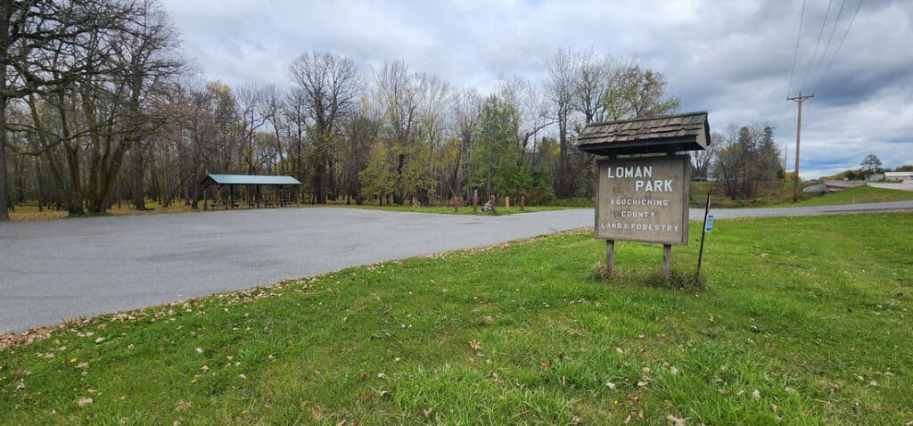 A wooden sign reads “Loman Park, Koochiching County Land/Forestry” next to a grassy area with a paved lot, trees in the background, and a covered picnic shelter. The sky is overcast. | Rainy Lake, MN