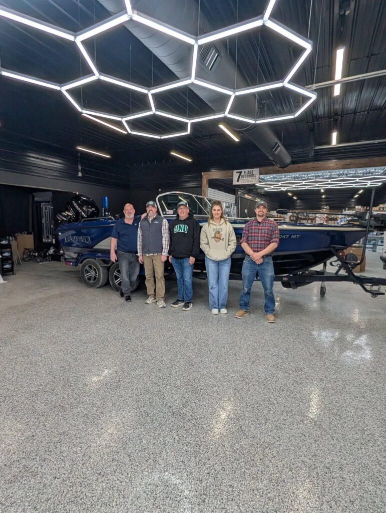Five people stand side by side in front of a blue boat inside the modern 7 Flags Outdoors showroom, featuring geometric ceiling lights and polished floors. | Rainy Lake, MN