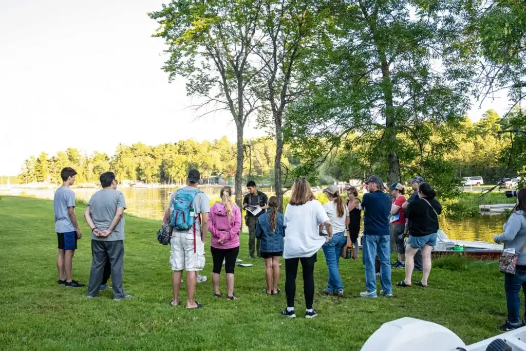 A group of people stands in a circle on a grassy area by a lake, gathered for a Summer Social organized by the conservancy. Trees and water create a scenic backdrop, while several people appear to be casually dressed for warm weather. | Rainy Lake, MN
