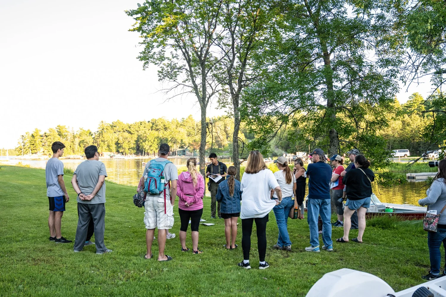 A group of people stands in a circle on a grassy area by a lake, gathered for a Summer Social organized by the conservancy. Trees and water create a scenic backdrop, while several people appear to be casually dressed for warm weather. | Rainy Lake, MN