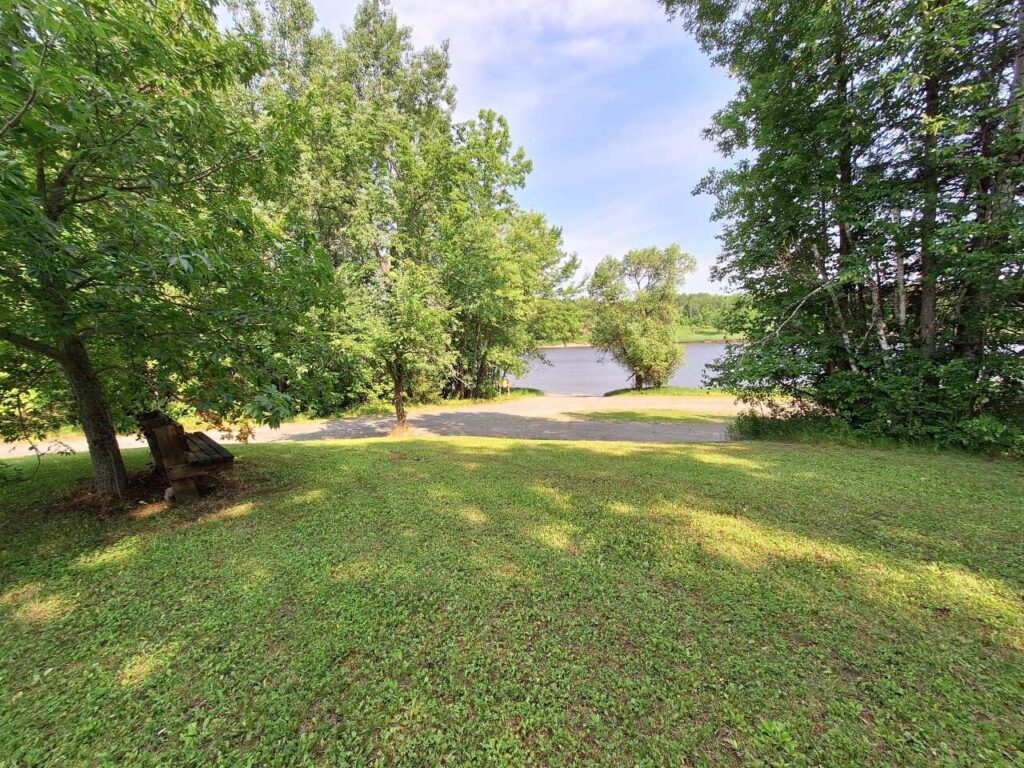 A grassy yard at Upper Sault Access is bordered by tall green trees and overlooks a calm river under a partly cloudy sky, with a dirt path running along the water’s edge. A wooden bench sits in the shade to the left. | Rainy Lake, MN