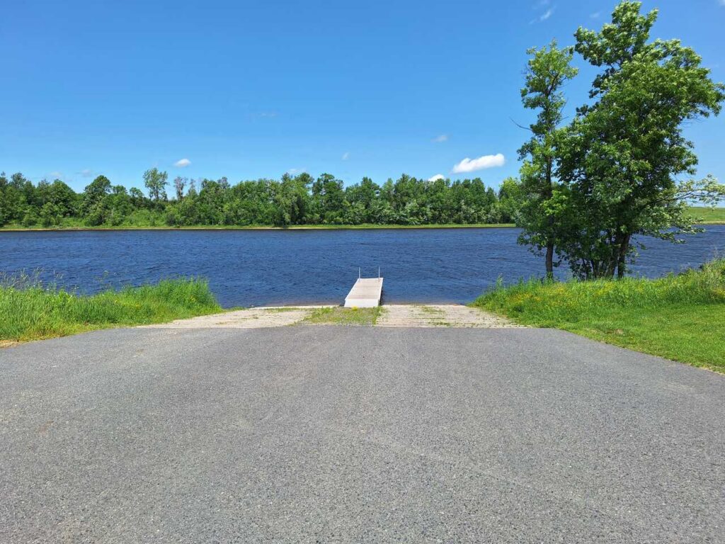 A paved boat ramp at Vidas Access leads to a small dock extending into a calm lake, surrounded by green grass and trees under a clear blue sky. | Rainy Lake, MN