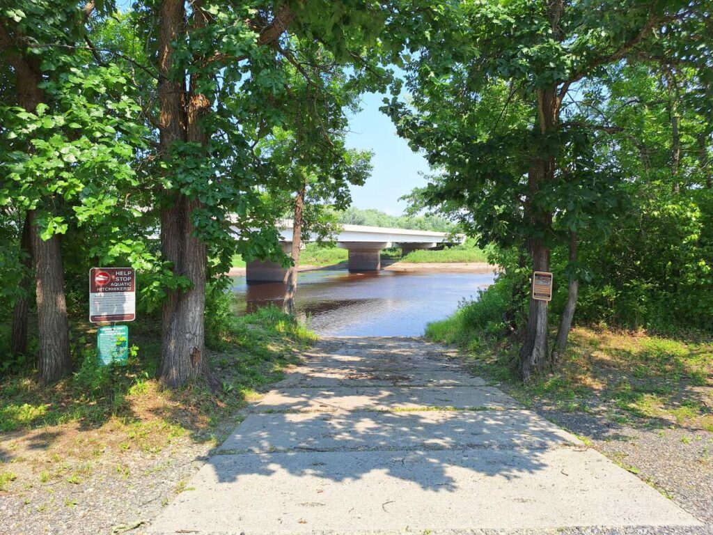 A concrete boat ramp leads to a river, bordered by trees on both sides. A bridge crosses the river in the background, and signs are posted on trees near the boat ramp. The scene is bright and sunny. | Rainy Lake, MN