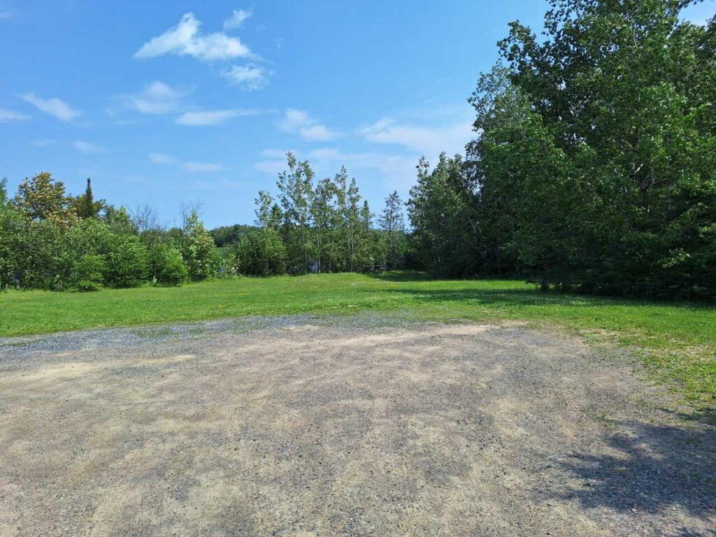 A sunny day at Wayside Park features a gravel area in the foreground, with green grass, trees, and a blue sky dotted with scattered clouds in the background. | Rainy Lake, MN
