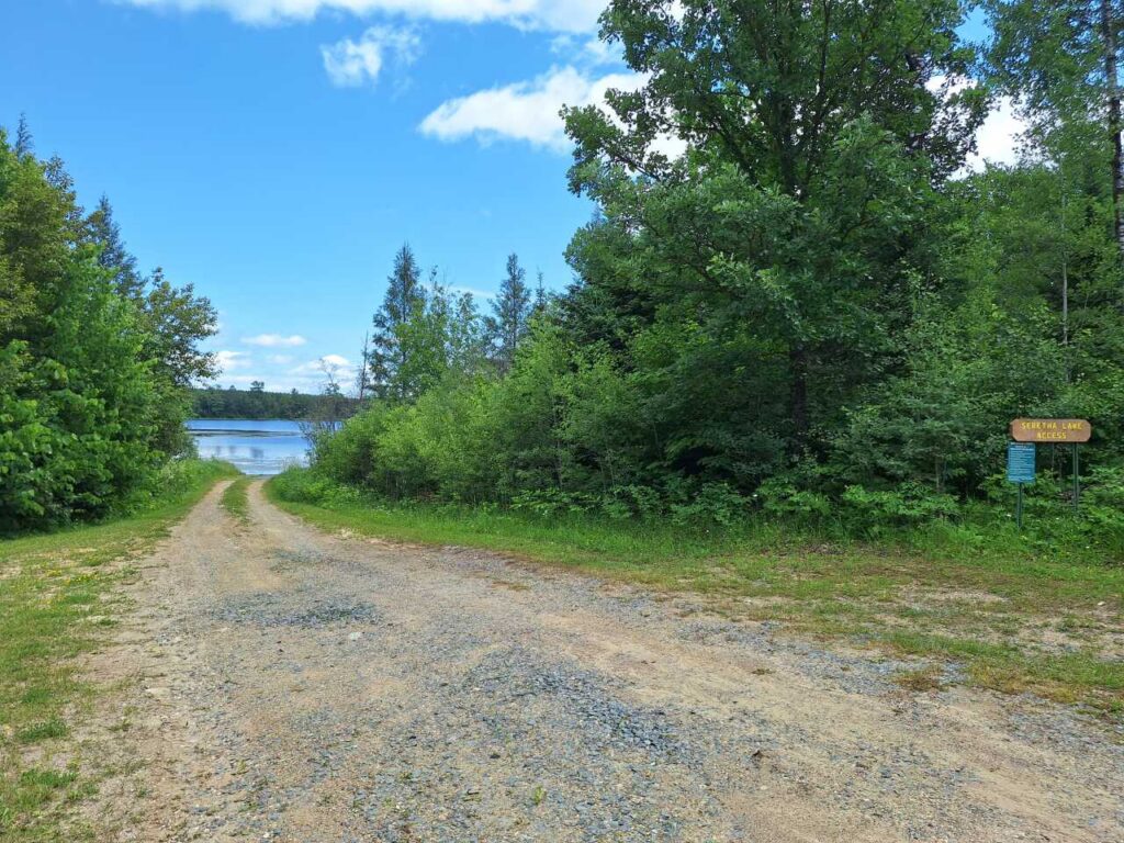 A gravel path leads through green trees toward Seretha Lake under a blue sky with scattered clouds. A wooden sign on the right side near the trees marks the Seretha Lake access. | Rainy Lake, MN