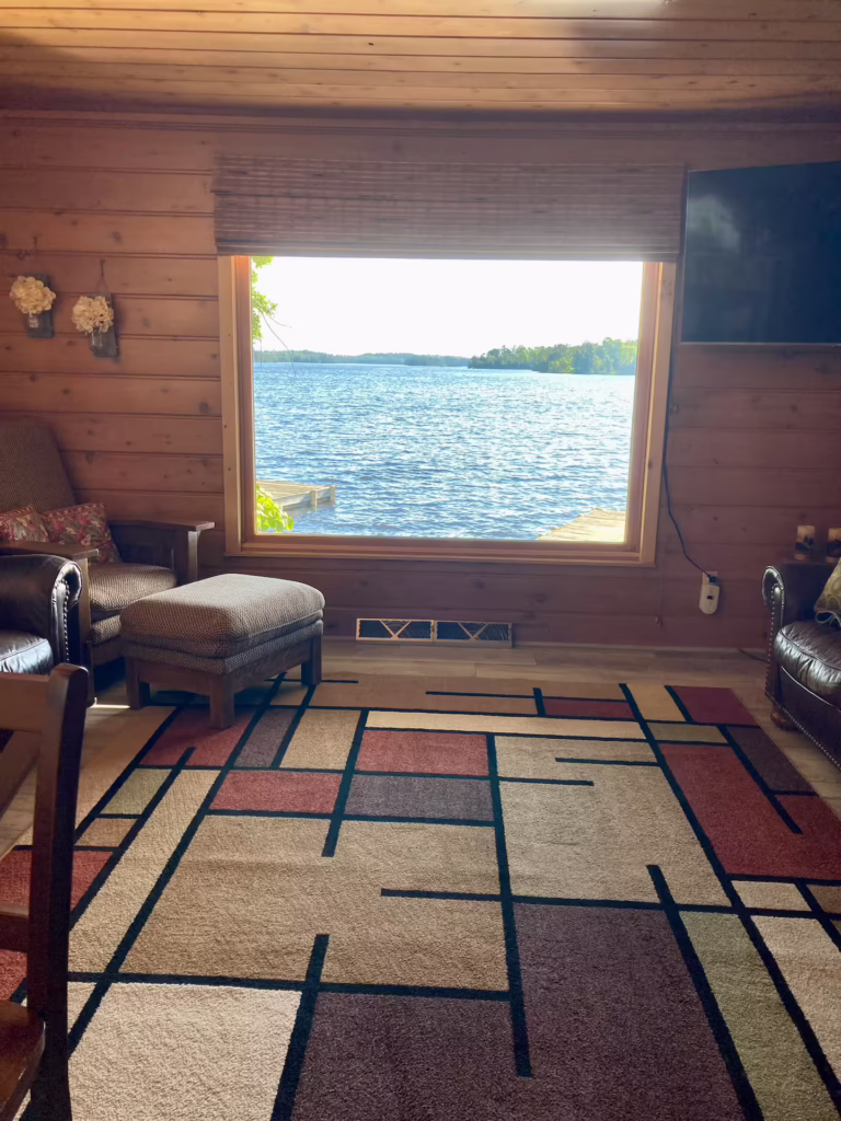 A cozy wooden cabin room with modern furniture and a large window offering a scenic view of Dove Island’s lake and trees outside under a bright sky—perfect for travel enthusiasts seeking relaxing island adventures. | Rainy Lake, MN