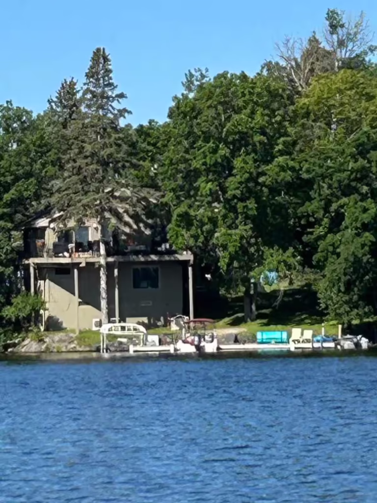 A house surrounded by trees sits beside a lake on beautiful Dove Island. Several boats are docked along the shoreline in front of the house under a clear blue sky, inviting you to experience unforgettable island travel and adventure tours. | Rainy Lake, MN