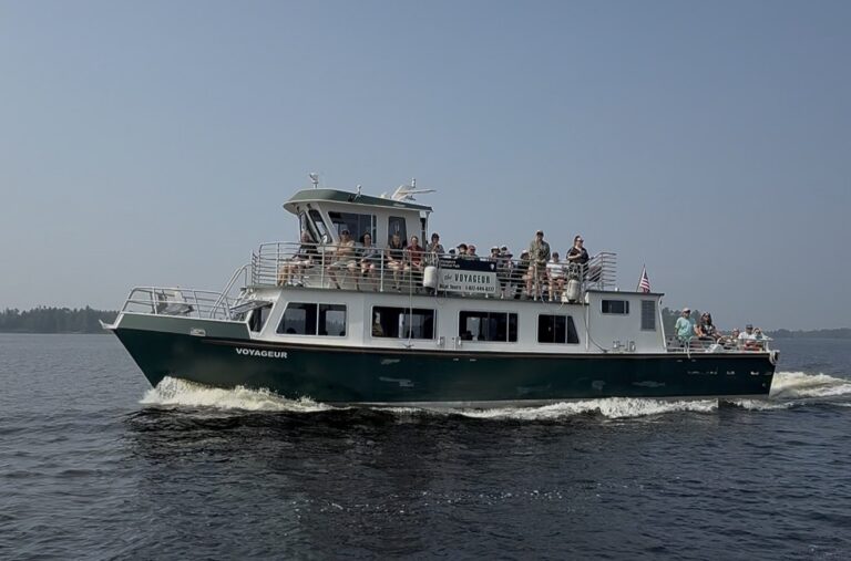 A green and white tour boat named “Voyageur” cruises on Rainy Lake, carrying passengers on the upper and lower decks. Trees are visible in the distant background under a hazy sky, creating a picturesque scene in Voyageurs National Park. | Rainy Lake, MN