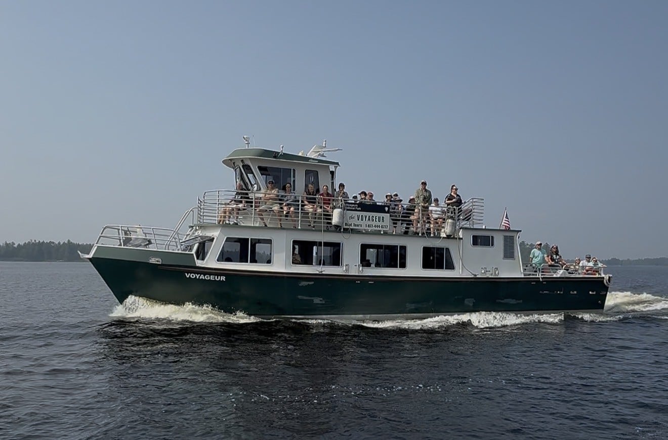 A green and white tour boat named “Voyageur” cruises on Rainy Lake, carrying passengers on the upper and lower decks. Trees are visible in the distant background under a hazy sky, creating a picturesque scene in Voyageurs National Park. | Rainy Lake, MN
