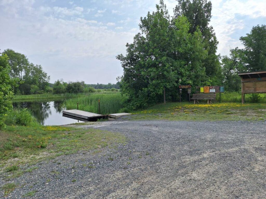 A small gravel parking area by a dock and the calm waters of the Rat Root River, surrounded by green trees and grass, with informational signs and a picnic table nearby at Watson Landing. | Rainy Lake, MN