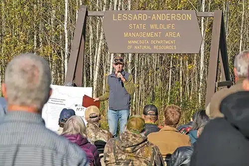 A person speaks to a seated outdoor audience near a sign reading “Lessard-Anderson State Wildlife Management Area, Minnesota Department of Natural Resources,” with trees in the background, highlighting the importance of wildlife conservation within this unique Wildlife Management Area. | Rainy Lake, MN