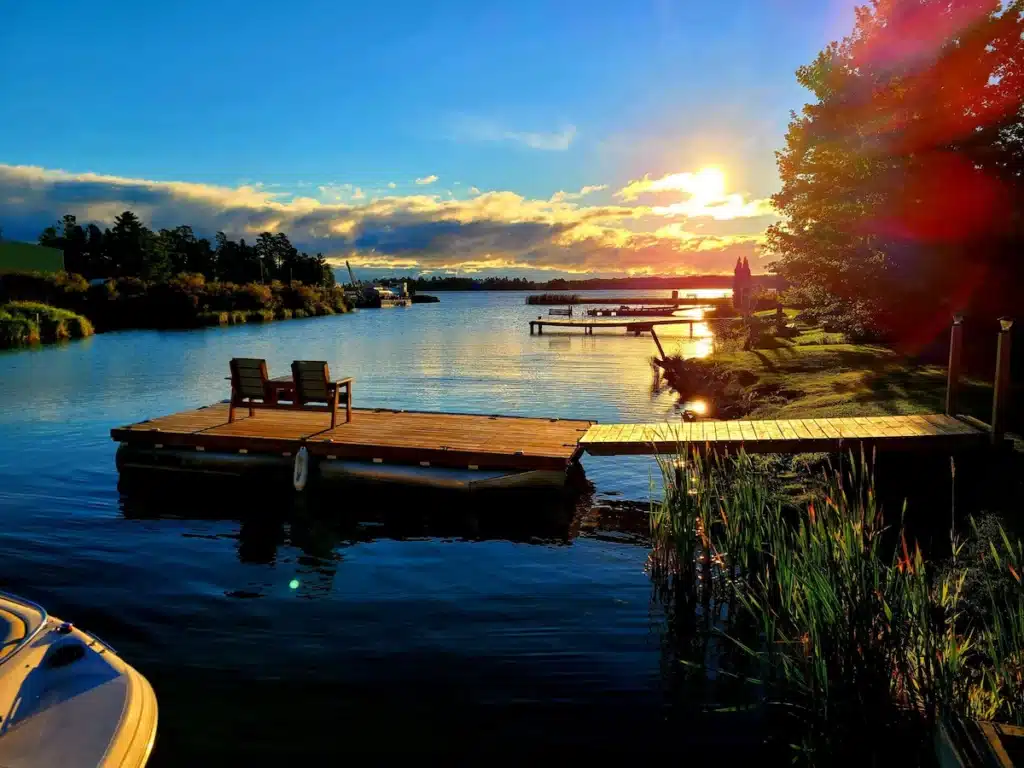 Two wooden chairs sit on a dock over a calm lake at sunset, evoking the peaceful ambiance of a Rainy Lake fishing outpost. The sun is low, casting golden light on the water and trees, while boats and distant docks dot the serene wilderness backdrop. | Rainy Lake, MN