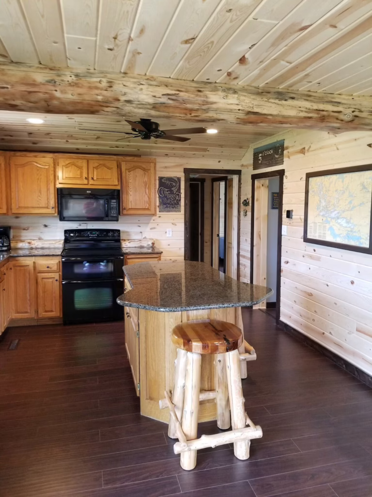 A rustic kitchen at the Rainy Lake Outpost features wooden cabinets, a granite island with a log-style stool, dark appliances, and light wood-paneled walls and ceiling. Brown wood flooring adds warmth, while doorways and cozy decor are visible in the background. | Rainy Lake, MN
