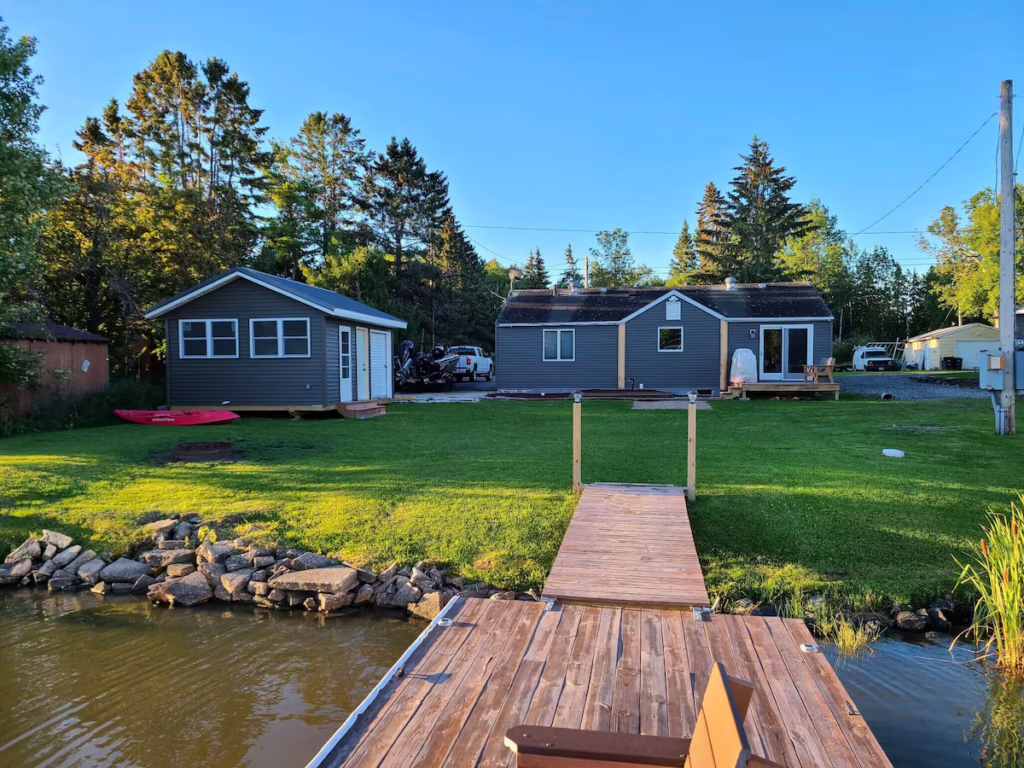 A wooden dock extends over Rainy Lake toward two gray outpost cabins with white trim, set on a green lawn surrounded by trees under a clear blue sky—an inviting setting for fishing and relaxation. | Rainy Lake, MN